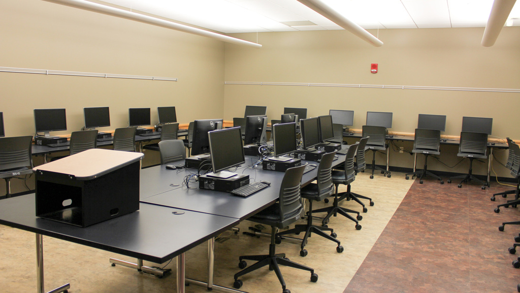 Image of computer classroom with computers sitting on tables in the middle and perimeter of room